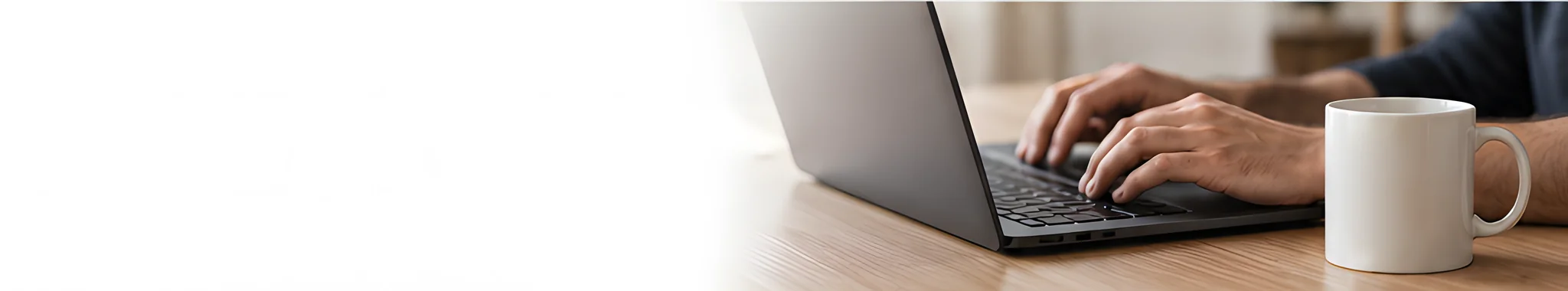 Hands typing on a laptop at a wooden desk beside a white mug