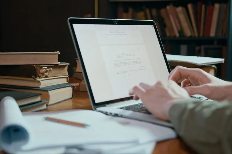 A writer types a draft on a laptop at a wooden desk, with stacked books, a pencil and printed notes beside it